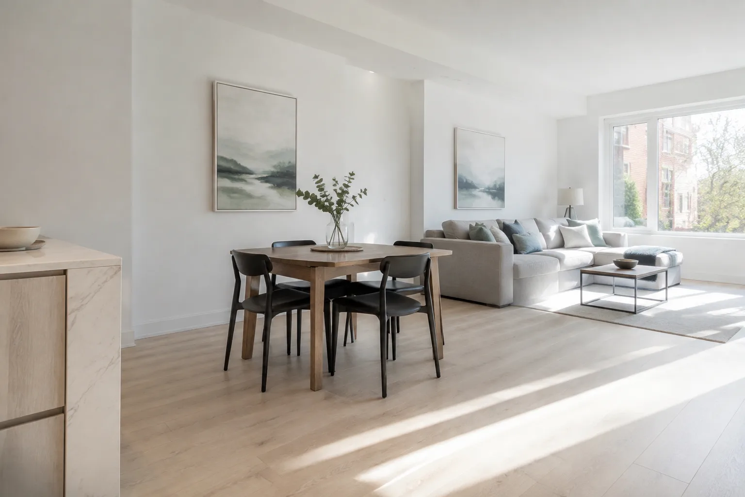 An open-concept living and dining room with a wood dining table, black chairs, a grey sectional sofa and framed landscape art, lit by a large window